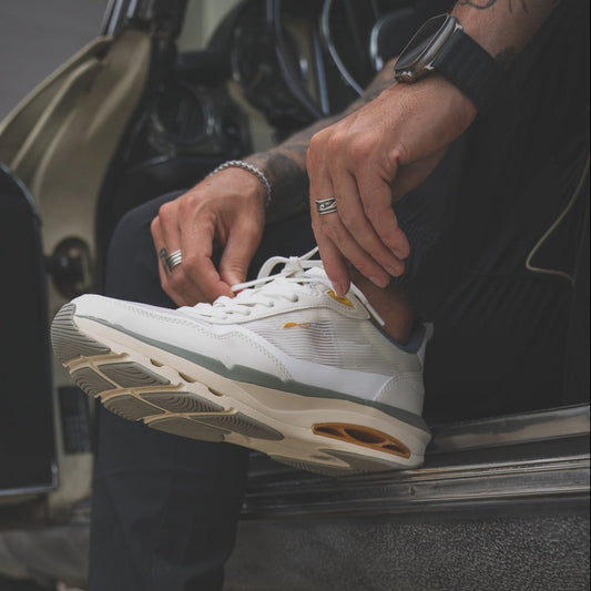 Man seated in an open car doorway lacing up a white CDNCE FRQNT Flyers sneaker