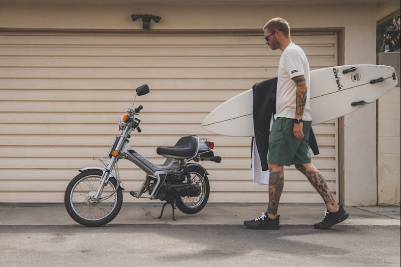 Man walking with a surfboard next to an electric bike in front of a garage.