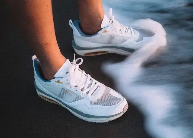 Off white sneakers with white soles worn by a person standing on a dark surface with water reflection.